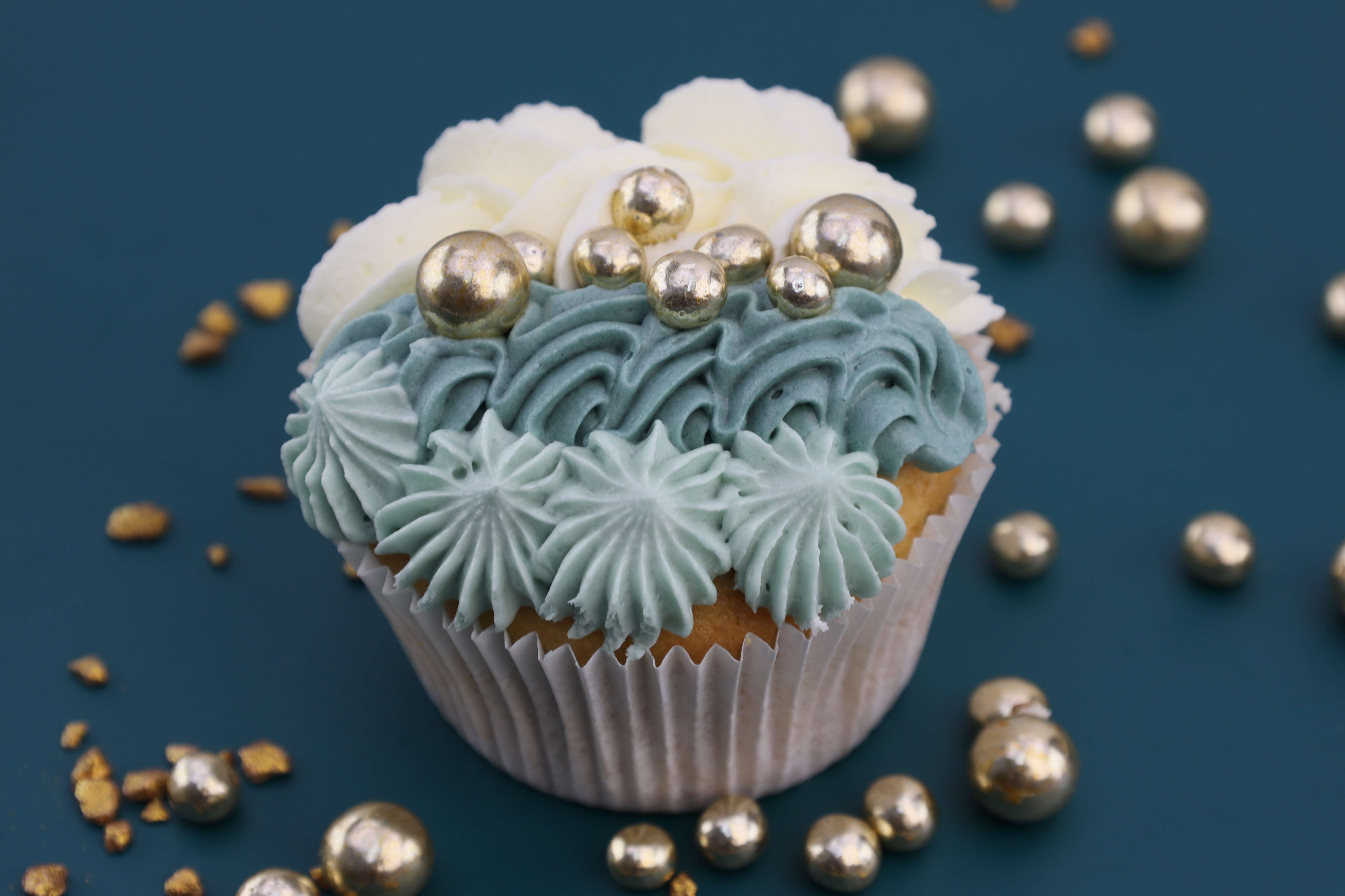 Köstlicher Muffins aus eukalyptusblauer und weißer Buttercreme mit goldenen essbaren Perlen oben drauf. 